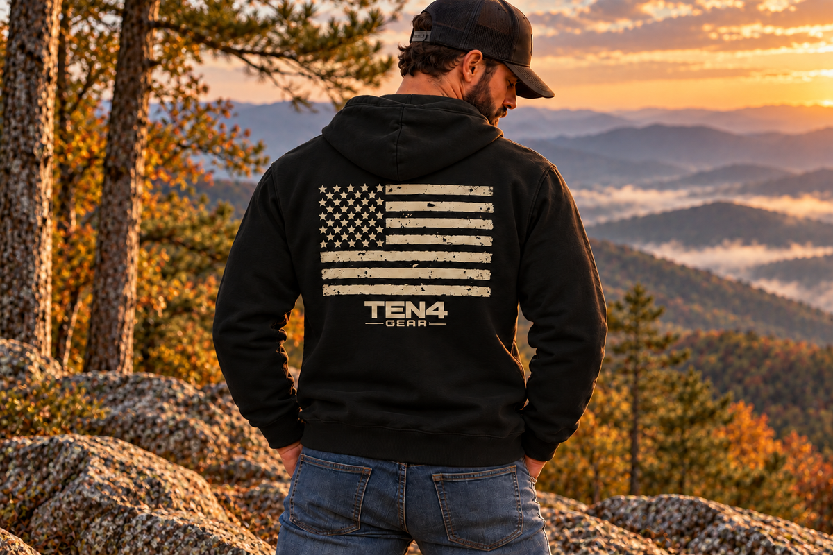 Black TEN4 American Pride hoodie with weathered tan flag and clean logo, on our bearded model at a wooded mountain overlook.