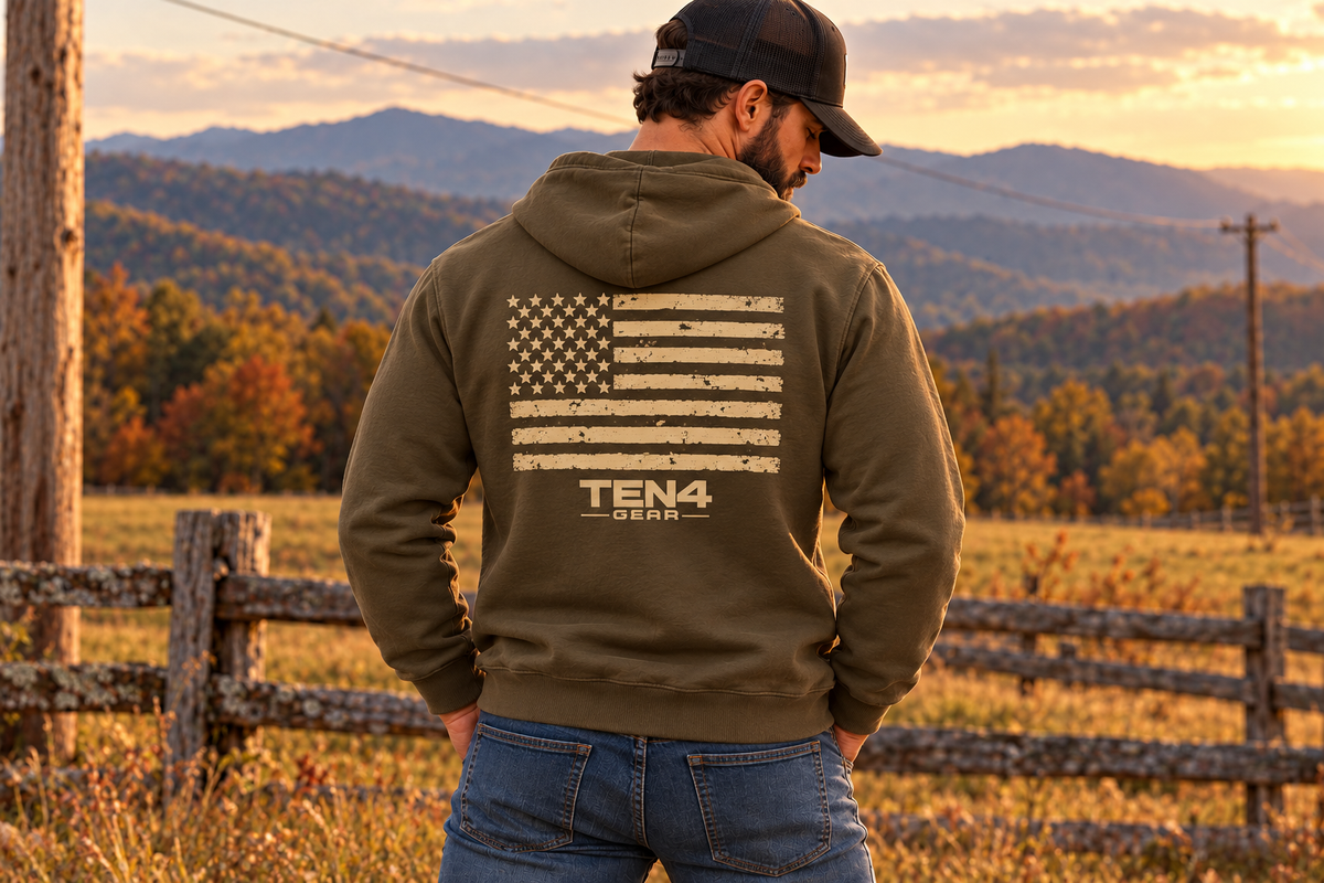 TEN4 American Pride Military Green hoodie, with a distressed tan flag and clean smaller TEN4 logo on the back, on our model at a rural mountain fence.
