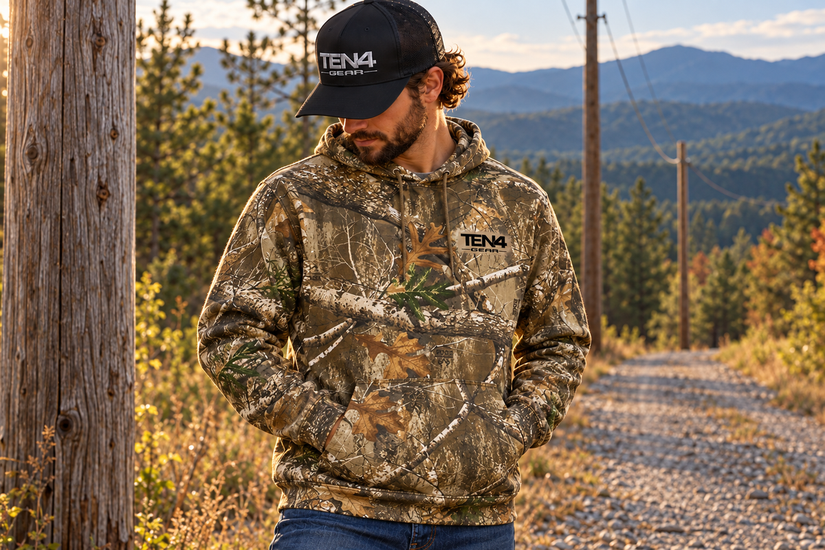 TEN4 Country Roots Hoodie — model in woodland camouflage beside wooden utility poles and mountains