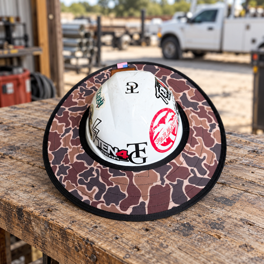 Duck Camo sunshade on a white hard hat resting on a rustic workbench beside a utility workshop