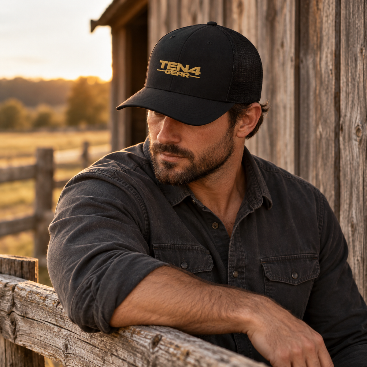 Black TEN4 full-logo trucker hat with Old Gold embroidery, shown on a model beside a rustic timber fence.