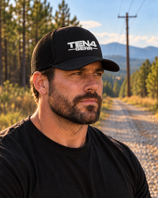 Black TEN4 Signature Trucker Hat with white embroidered TEN4 Gear logo, worn by a rugged man looking off-camera