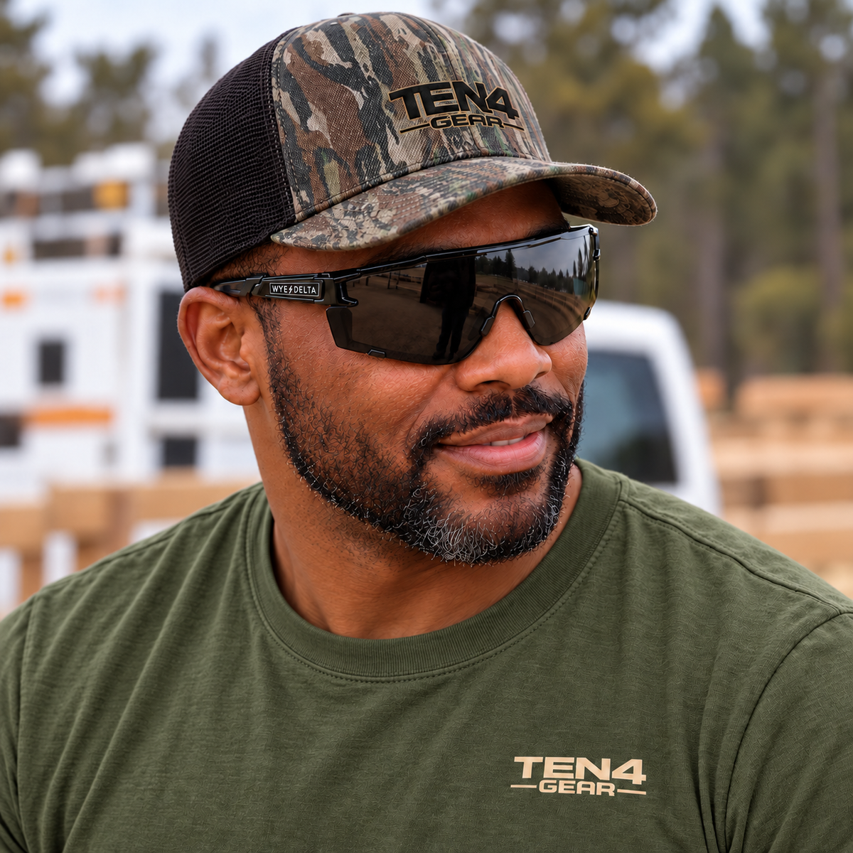 Lifestyle mockup of the TEN4 Full Logo Woods Camo/Black trucker hat, worn with black HiViz sunglasses and an olive TEN4 shirt.