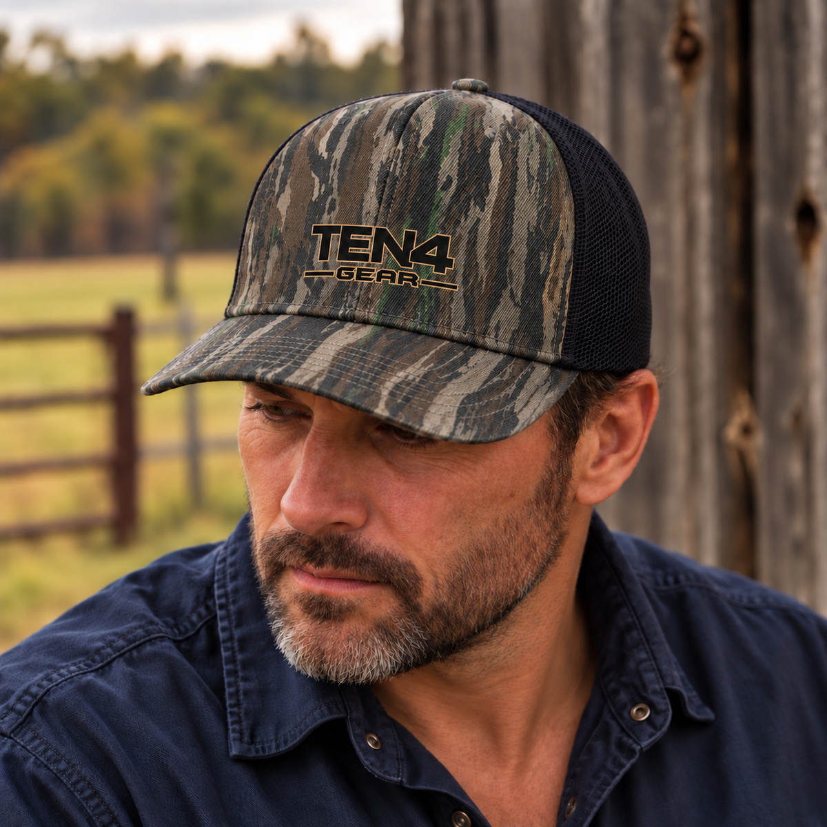 TEN4 woods camo trucker hat with a small black full logo and fine tan outline, worn by a man looking down beside a rustic barn.