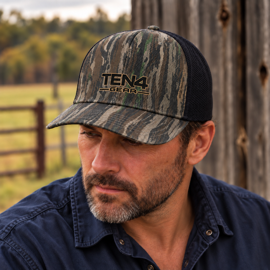 TEN4 woods camo trucker hat with a small black full logo and fine tan outline, worn by a man looking down beside a rustic barn.