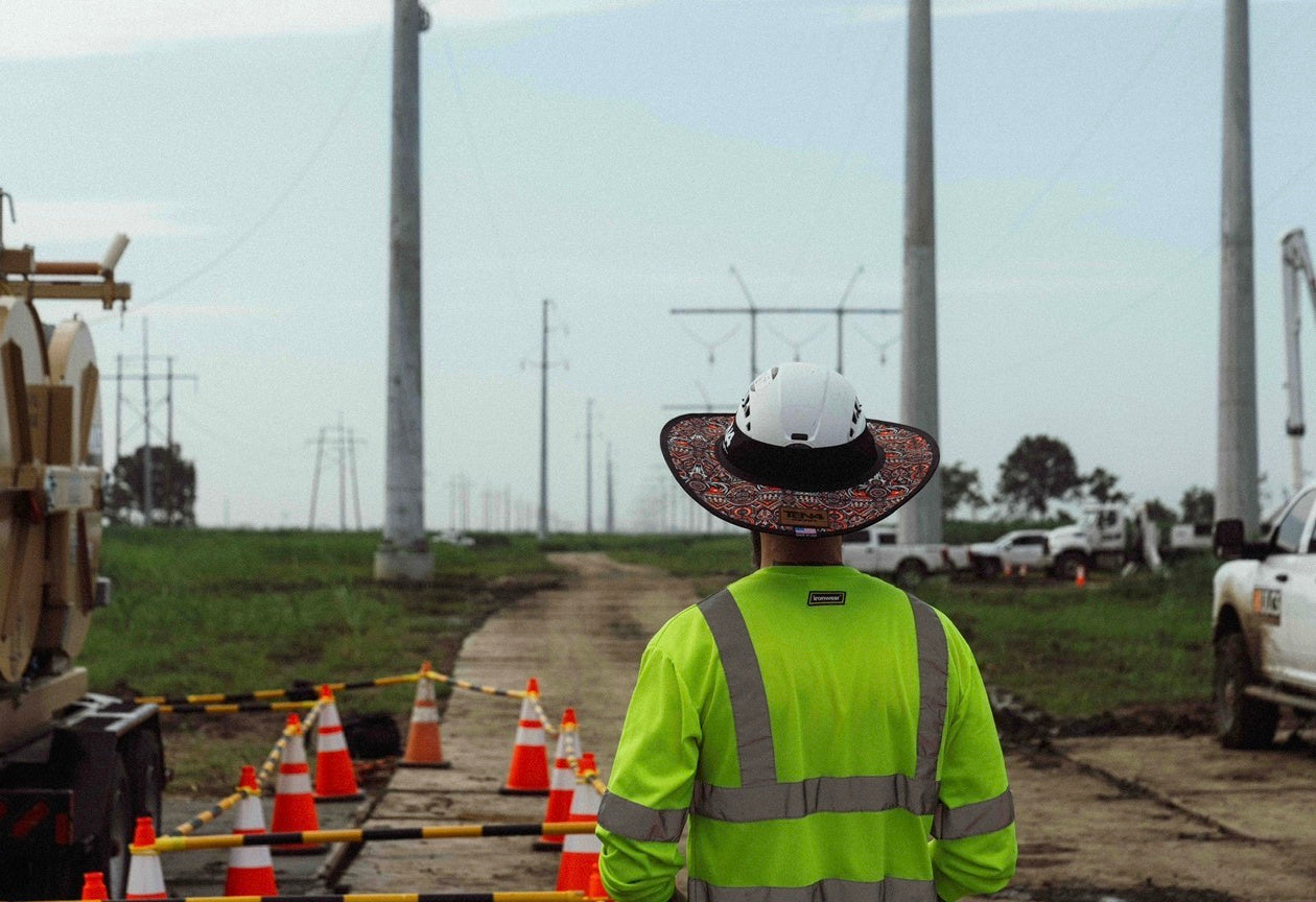 Lineman wearing a TEN4 patterned sunshade on a white hard hat at a transmission line jobsite, photographed from behind.